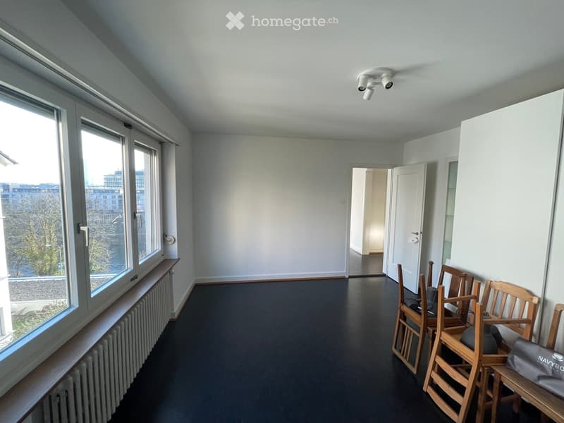 A room with large windows, a radiator, and several wooden chairs stacked against the wall.