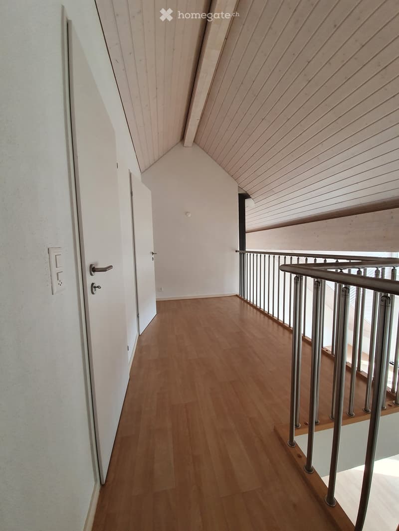 A narrow hallway with wooden flooring, white walls, and a modern metal staircase railing leading upwards.