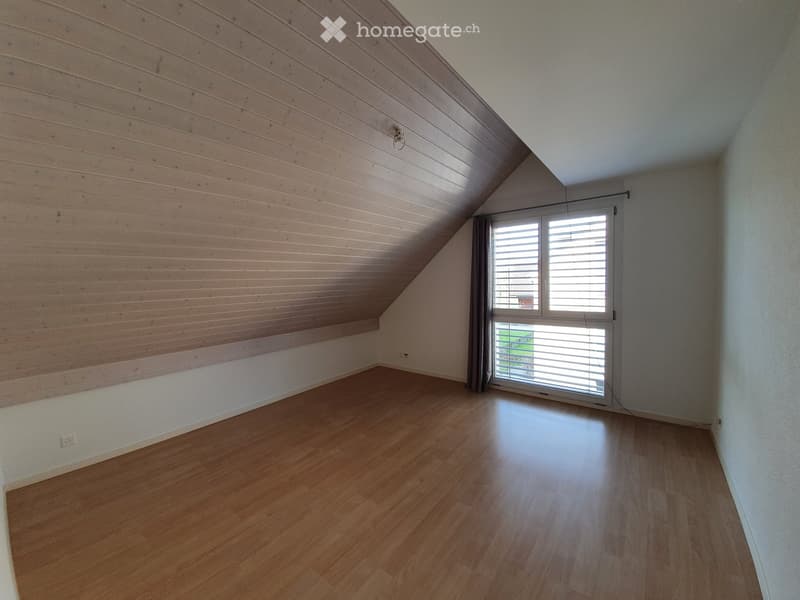 Empty attic room with wooden walls and floor, large window with blinds, and exposed wooden beams on the ceiling.