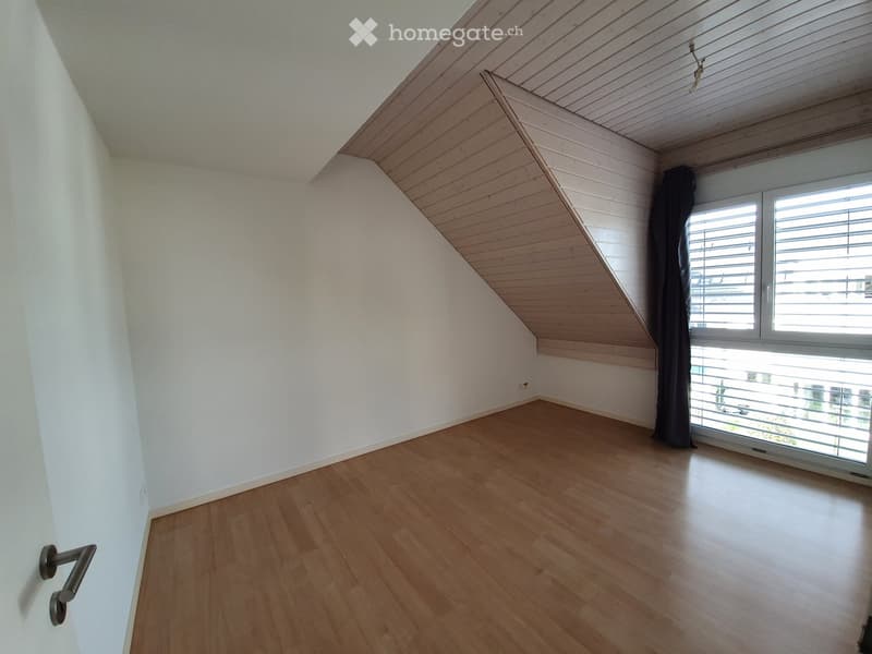 Empty room with wooden flooring, white walls, and a slanted wooden ceiling. Features a large window with blinds and a metal curtain rod.