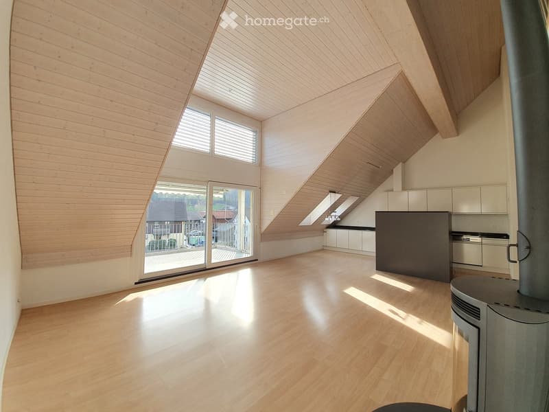 Open-plan attic space with wooden floors, slanted wooden ceiling, large windows, and a modern kitchen area with cabinets and a wood stove.