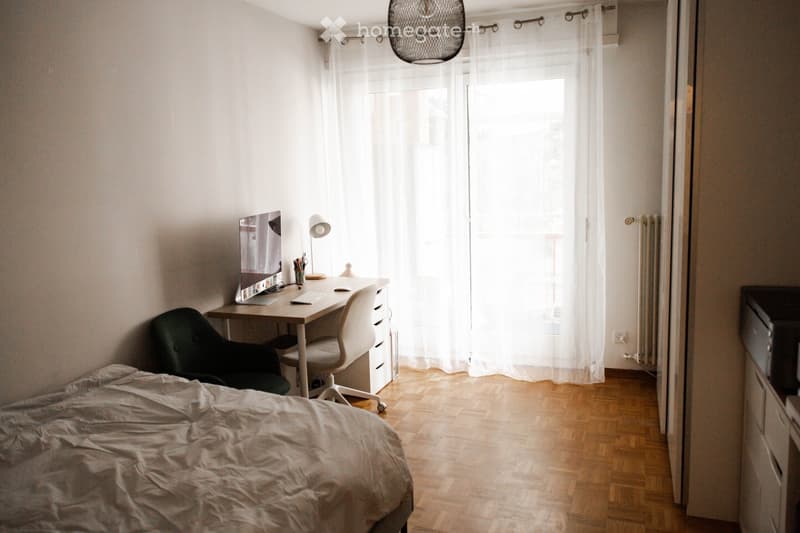 Cozy bedroom with a bed, desk, and chair. The room features wooden flooring, white walls, and large windows with sheer curtains.