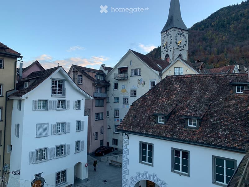 A picturesque European village scene with traditional multi-story buildings, a prominent clock tower, and a stone-paved square.