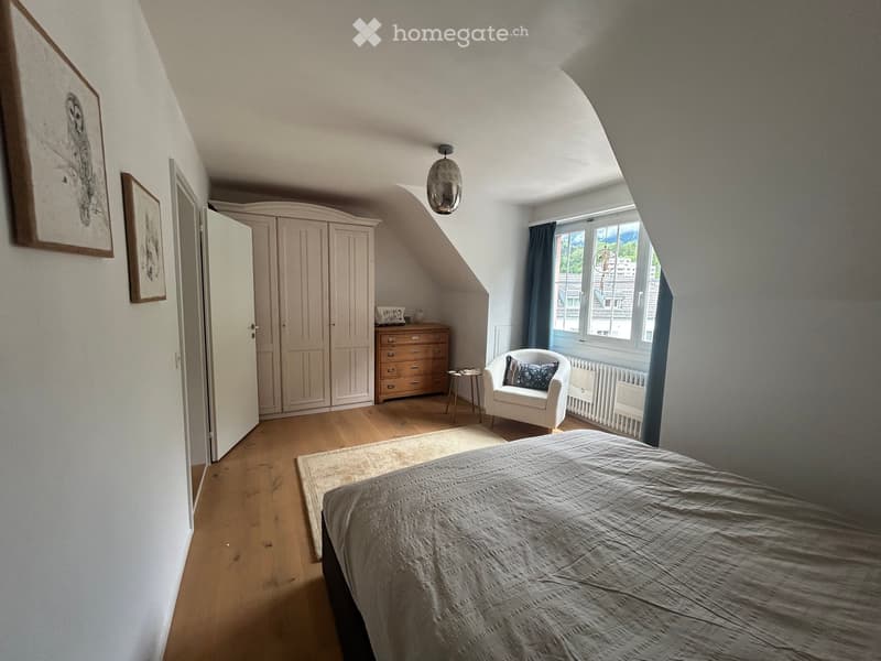 Cozy attic bedroom with wooden flooring, slanted ceilings, large window with blue curtains, bed with grey duvet, white armchair, small round table, built-in wardrobe, and framed artwork on the walls.