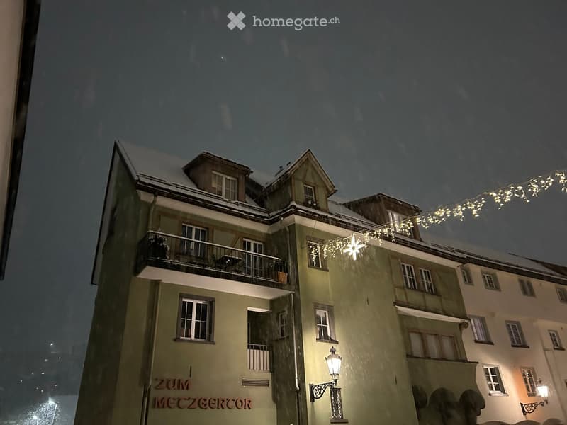 Multi-story building with snow-covered roof and balconies, illuminated by string lights and street lamps.