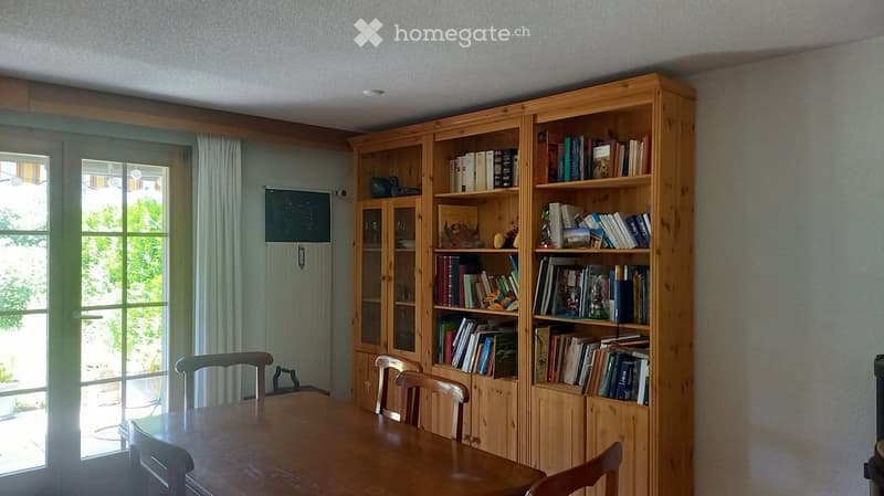 A well-lit dining area with a wooden table and chairs, a large wooden bookcase filled with books and decorative items, and large windows with curtains.