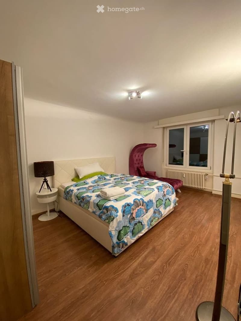 Cozy bedroom with wooden flooring, a large bed with a colorful duvet, a nightstand with a lamp, a red accent chair, and a radiator beneath a window.