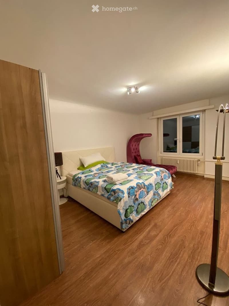Cozy bedroom with wooden flooring, a large bed with a colorful duvet, a nightstand with a lamp, a red chair, and a radiator under the window.