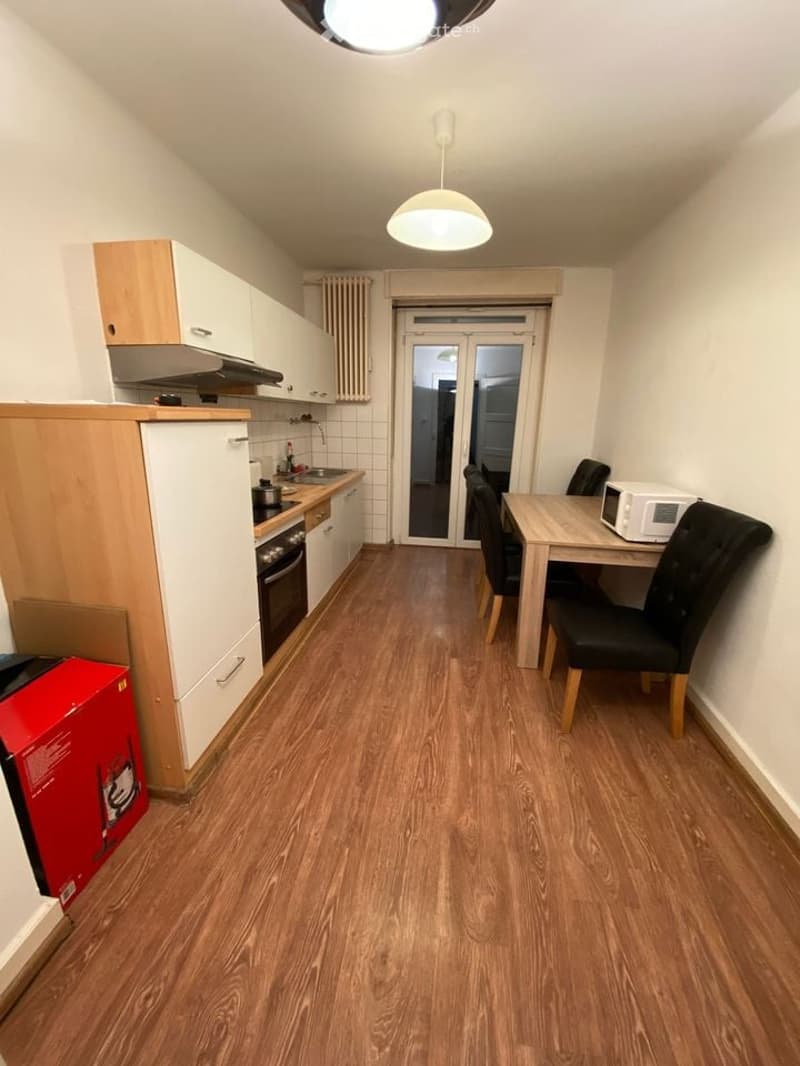 Compact kitchen area with light wood cabinets, a stove, sink, and a small dining table with chairs. The floor is wooden, and there is a sliding glass door leading to another room.