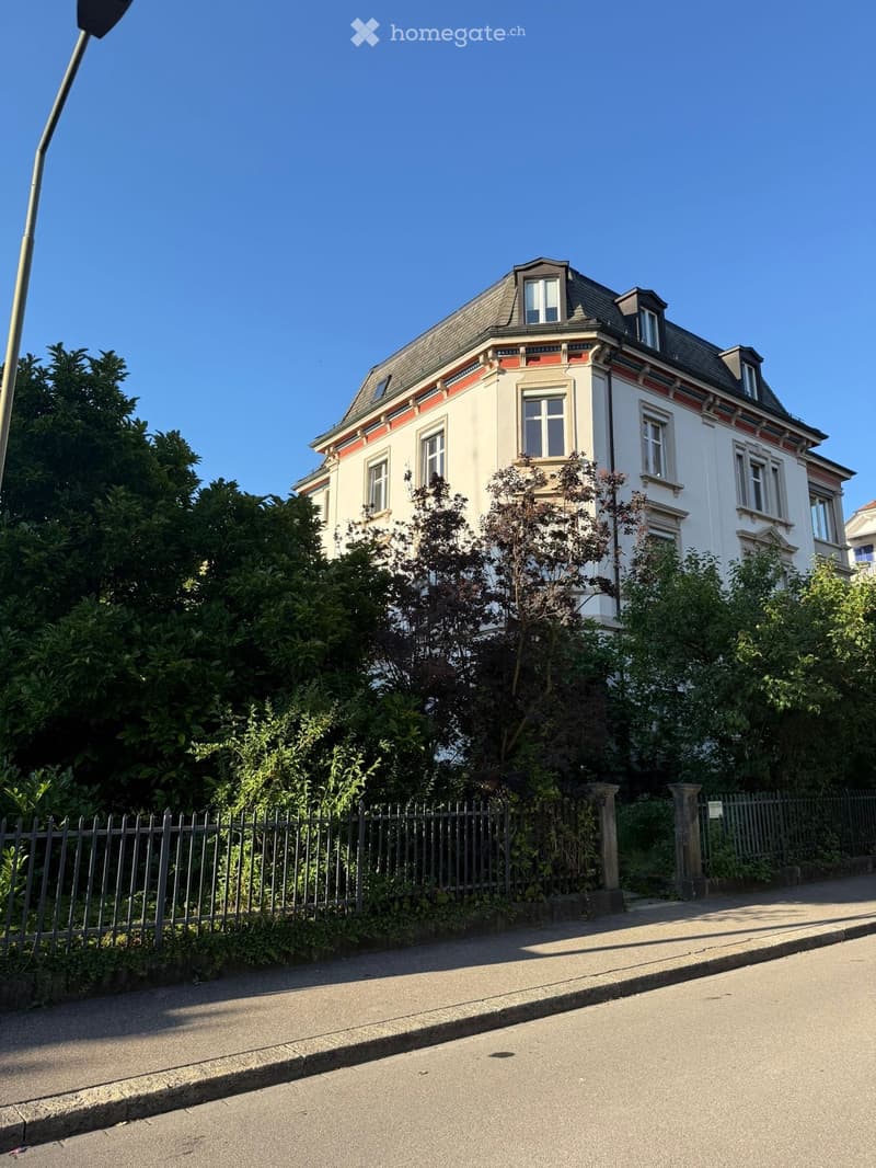 A two-story residential house with white walls, multiple windows, and a grey roof, surrounded by greenery and a metal fence.