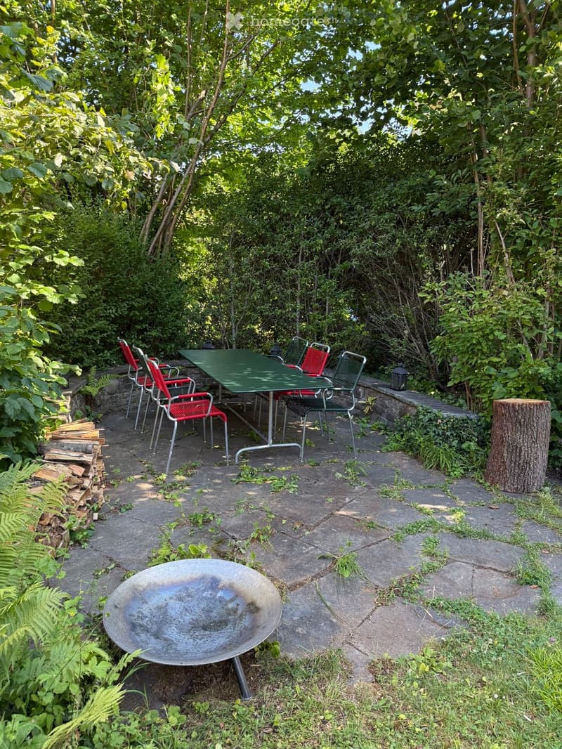 A lush, green garden patio with a stone floor, a green metal table surrounded by red and green chairs, a fire bowl, and a stack of firewood. The area is surrounded by dense foliage and trees.