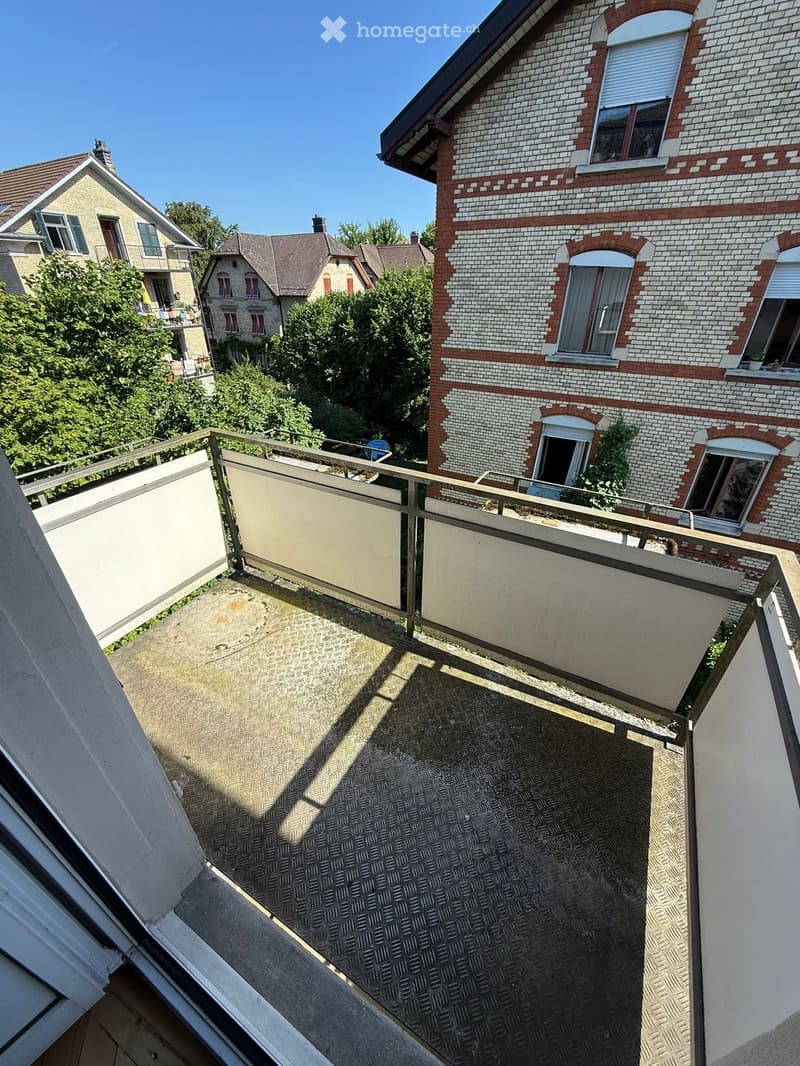 Balcony with metal railing, concrete floor, and view of neighboring houses and greenery.