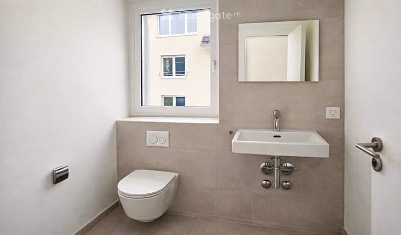 Modern bathroom with a toilet, sink, and mirror. Light-colored tiles and a window provide natural light.