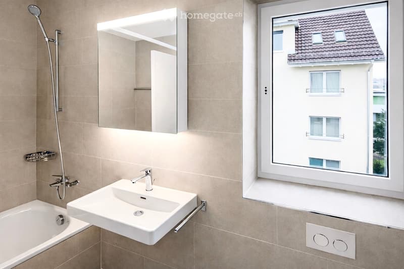 Modern bathroom with a bathtub, white sink, mirror, and showerhead. The room features beige tiles and a large window offering a view of a building exterior.