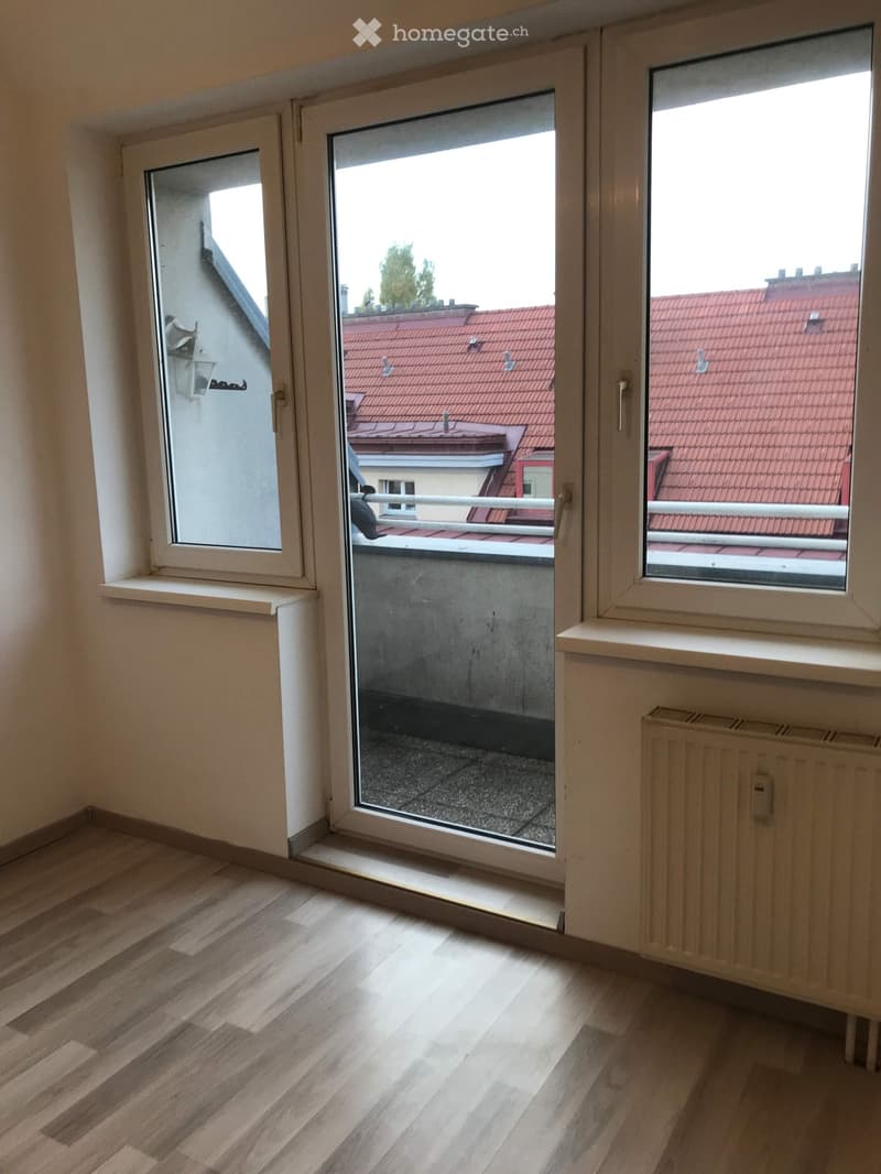 Empty room with wooden flooring, white walls, large windows, radiator, and a view of rooftops through the open window.