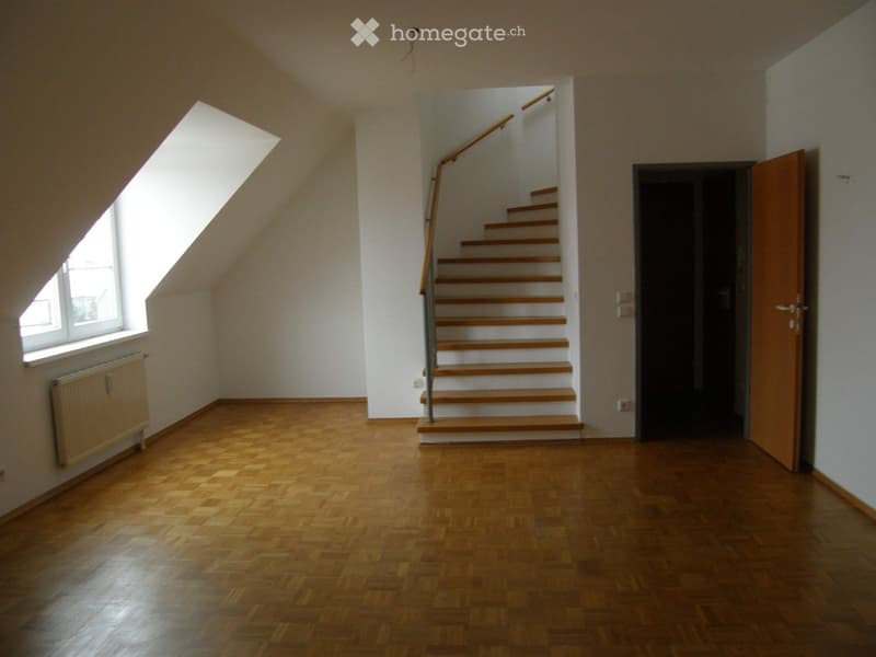 Empty room with wooden flooring, white walls, a staircase leading upwards, a window with a radiator below, and a closed wooden door.