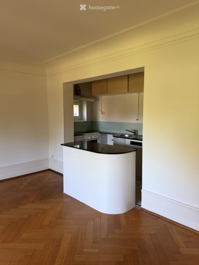 Open-plan kitchen and living area with light wood flooring, white cabinetry, and a central island. The kitchen features a stove, sink, and countertop, while the living area has a large window and neutral-colored walls.
