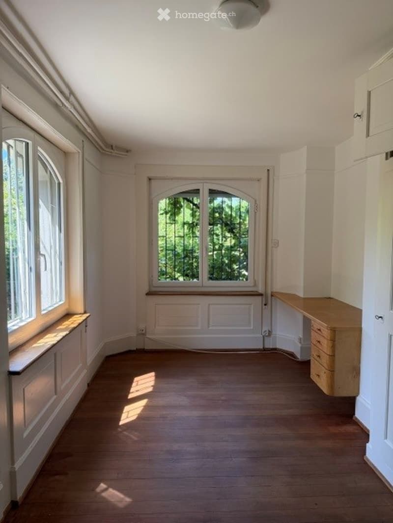 Empty room with wooden flooring, white walls, large windows, and a small wooden desk in the corner.