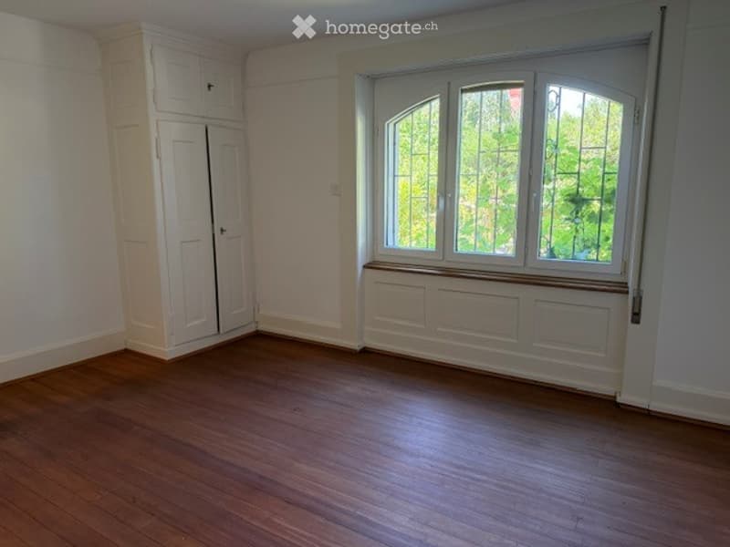 Empty room with wooden flooring, white walls, large window with multiple panes, built-in white cabinetry, and a radiator cover below the window.