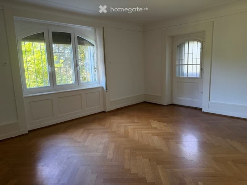 Empty room with wooden parquet flooring, large windows with white frames, and a white door.