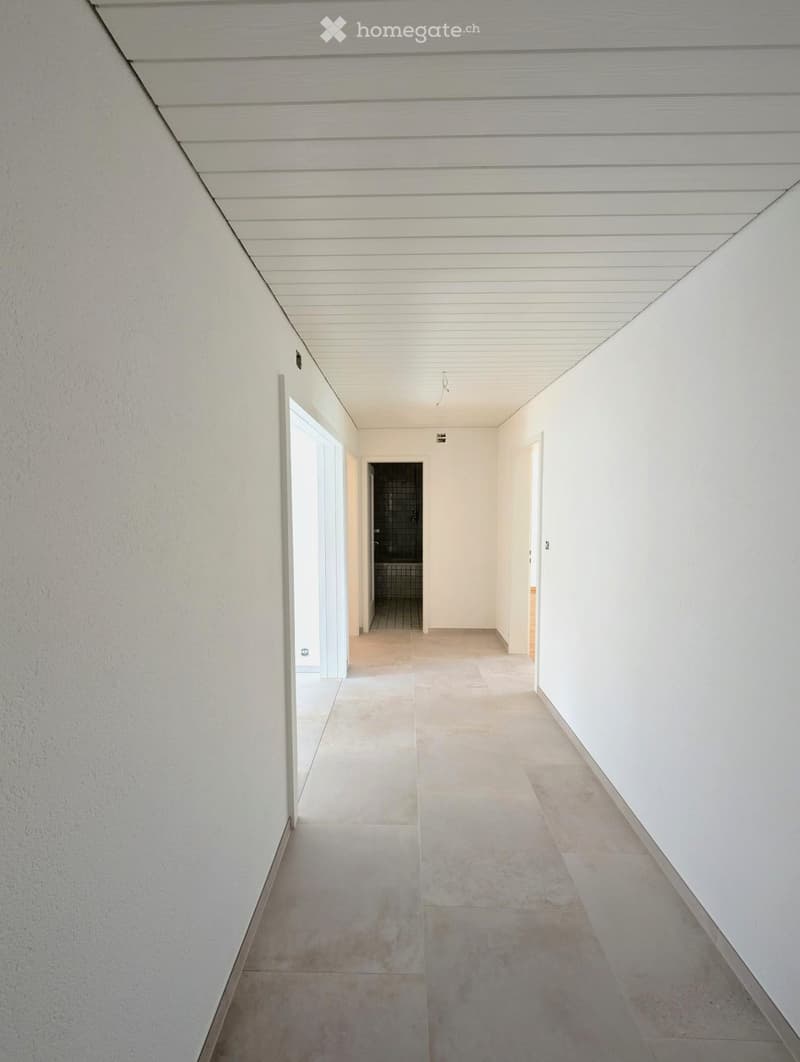 A long, narrow hallway with white walls, a white paneled ceiling, and beige tiled flooring. The hallway features several doors and a visible bathroom at the end.