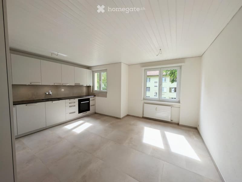 Modern kitchen with white cabinets, stainless steel sink, oven, and tiled floor. Large windows provide natural light.