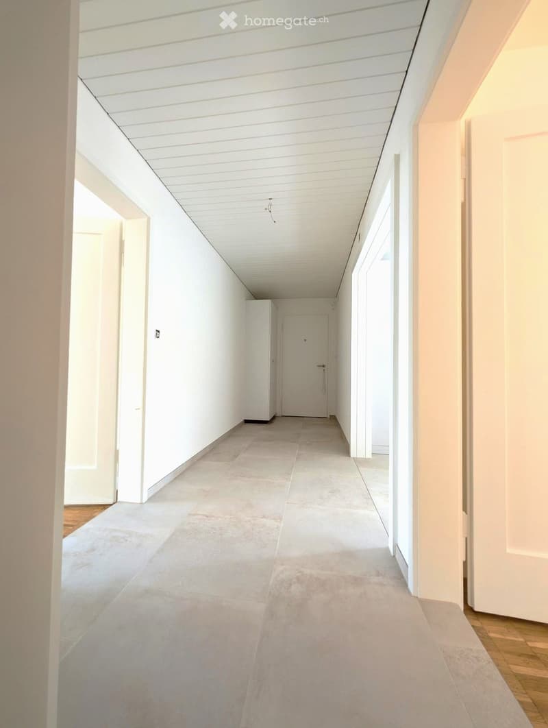 Modern, minimalist hallway with white walls, grey tiled floor, and white ceiling. Features several doors and a small niche on the left side.