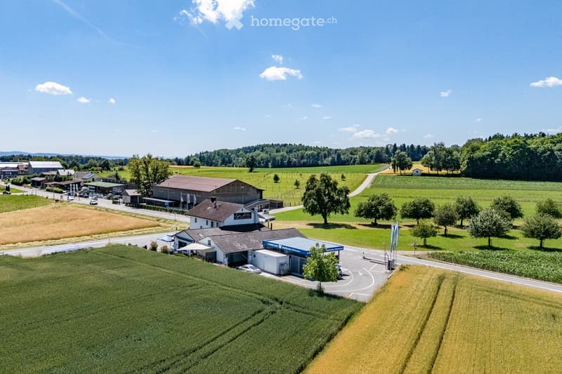 Aerial view of a rural building complex with a large main building, smaller outbuildings, parking area, and surrounding farmland.