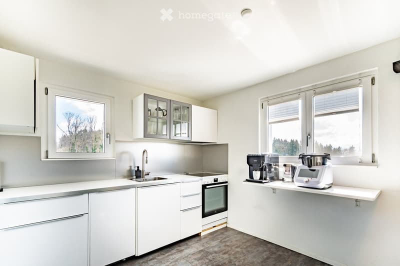 Modern kitchen with white cabinets, stainless steel appliances, and a breakfast bar with coffee maker and blender. Large windows provide natural light.