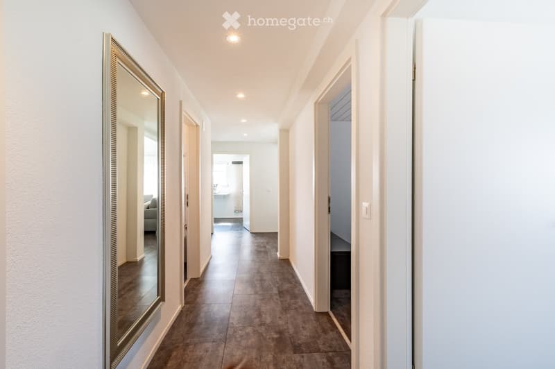 A long, well-lit hallway with beige walls, dark tiled flooring, and several open doors leading to adjacent rooms. A large mirror is mounted on one wall, and recessed ceiling lights provide illumination.