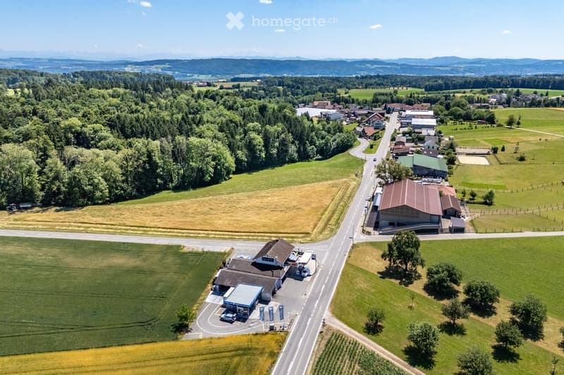 Aerial view of a rural area with a gas station, a large barn, scattered houses, and surrounding farmland and forests.