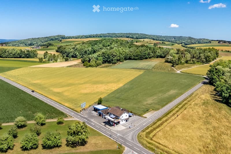 Aerial view of a rural area featuring a small gas station with a blue canopy, surrounded by agricultural fields and a road passing through the landscape.