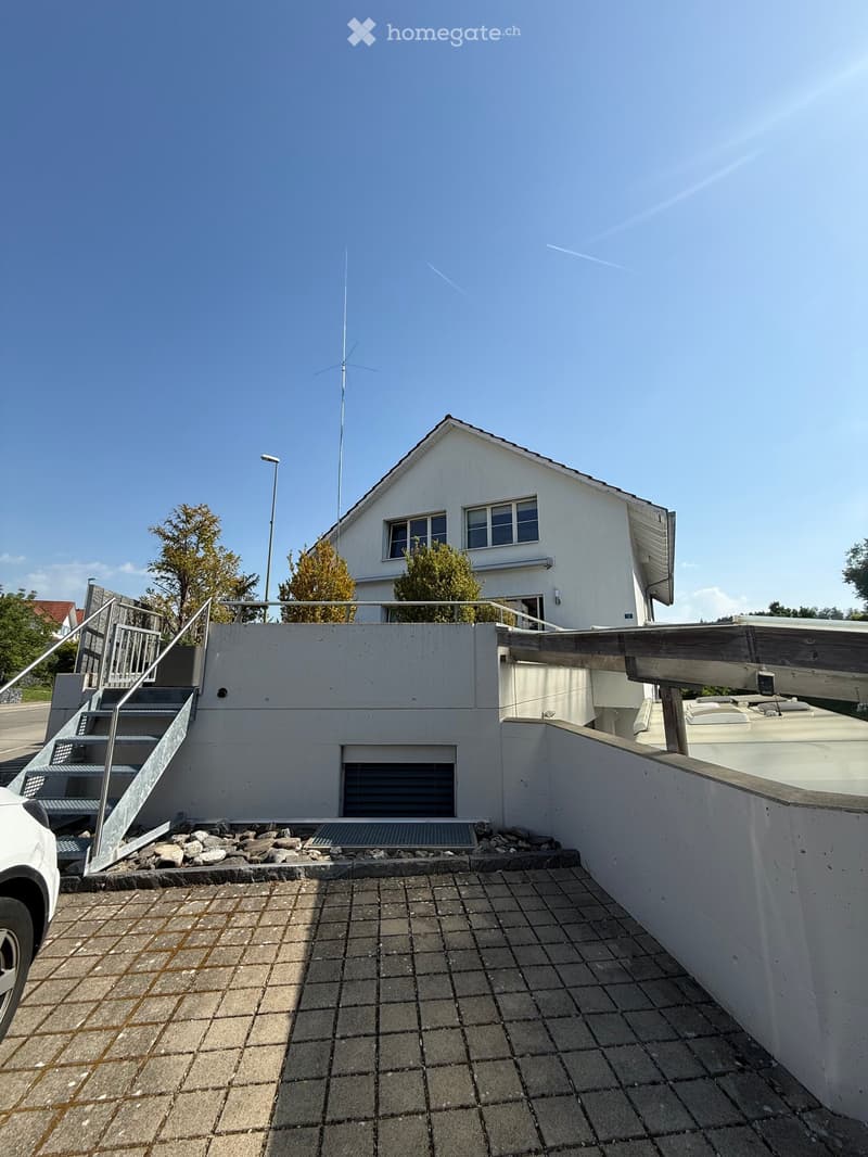 Two-story white house with a raised ground floor, metal staircase, and antenna on the roof.
