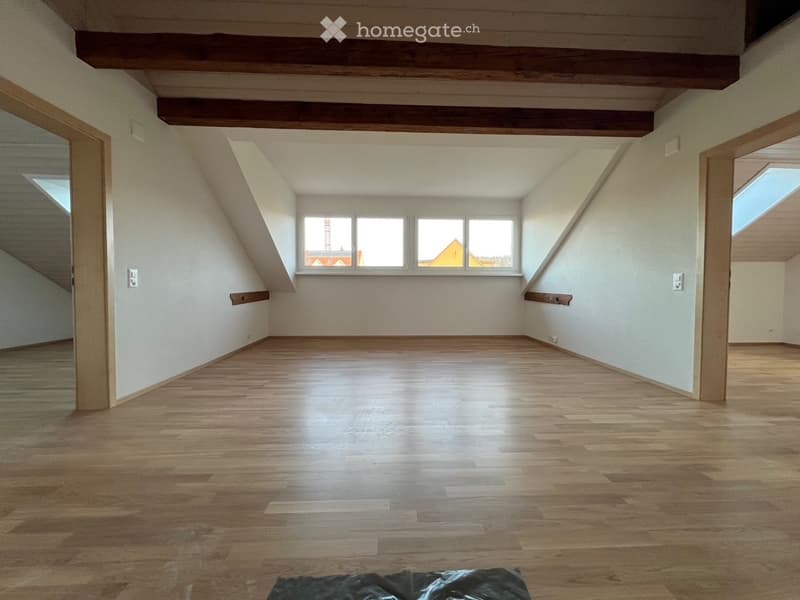 Empty attic room with wooden floors, exposed wooden beams, and large windows letting in natural light.