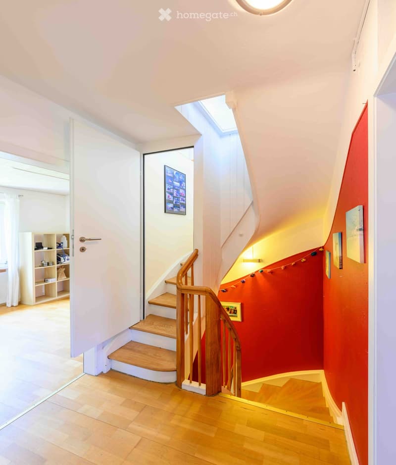 Spacious hallway with wooden flooring, a staircase with wooden railings, and colorful wall decorations. Features a skylight and a small shelf with items on the left side.