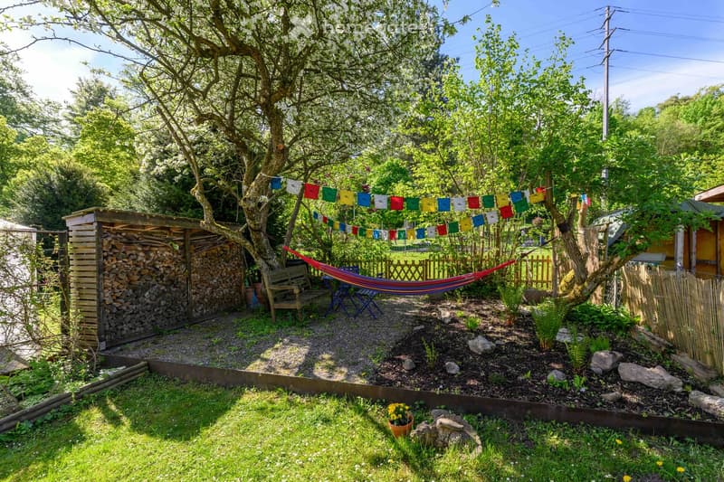 A serene garden area with a hammock, wooden bench, colorful prayer flags, a stack of firewood, and lush greenery.