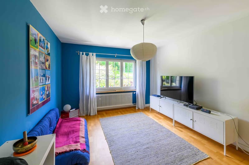 Living room with blue walls, wooden floor, sofa with pink blanket, TV on white cabinet, round pendant light, and a small table with a decorative bowl.