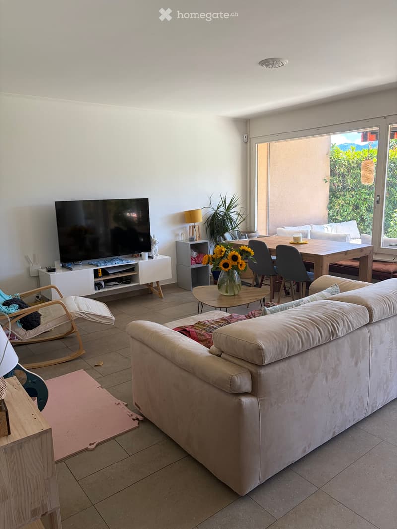 Modern living room with a beige sofa, coffee table, dining table, chairs, TV, and a vase with sunflowers. Large windows offer a view of greenery outside.