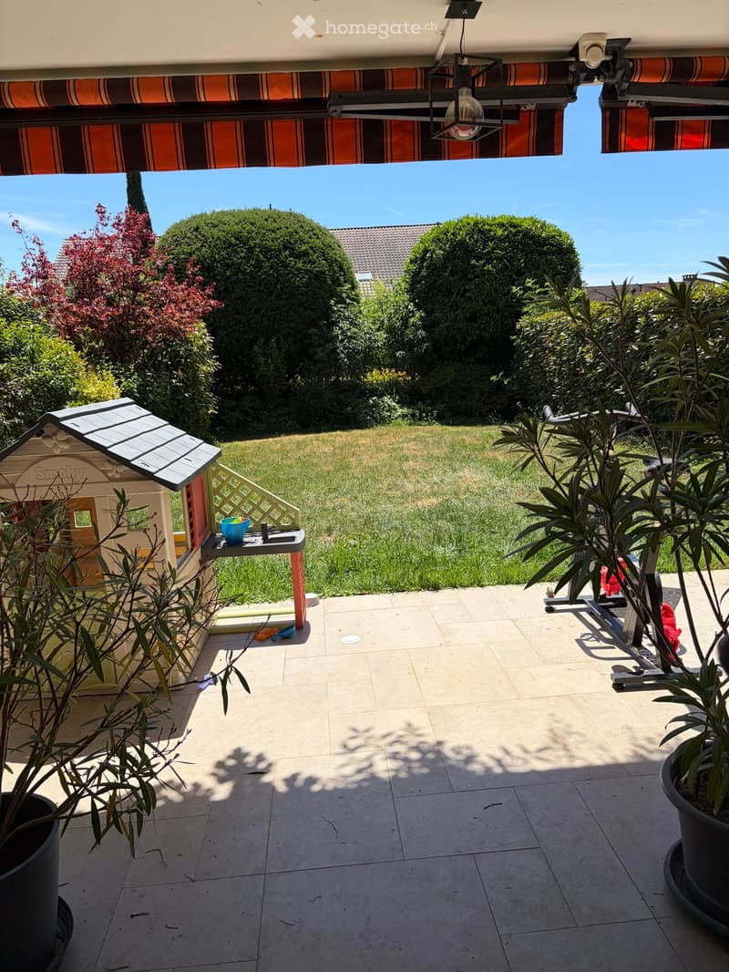 A sunny patio area with tiled flooring, potted plants, a small playhouse, and a grassy lawn in the background.