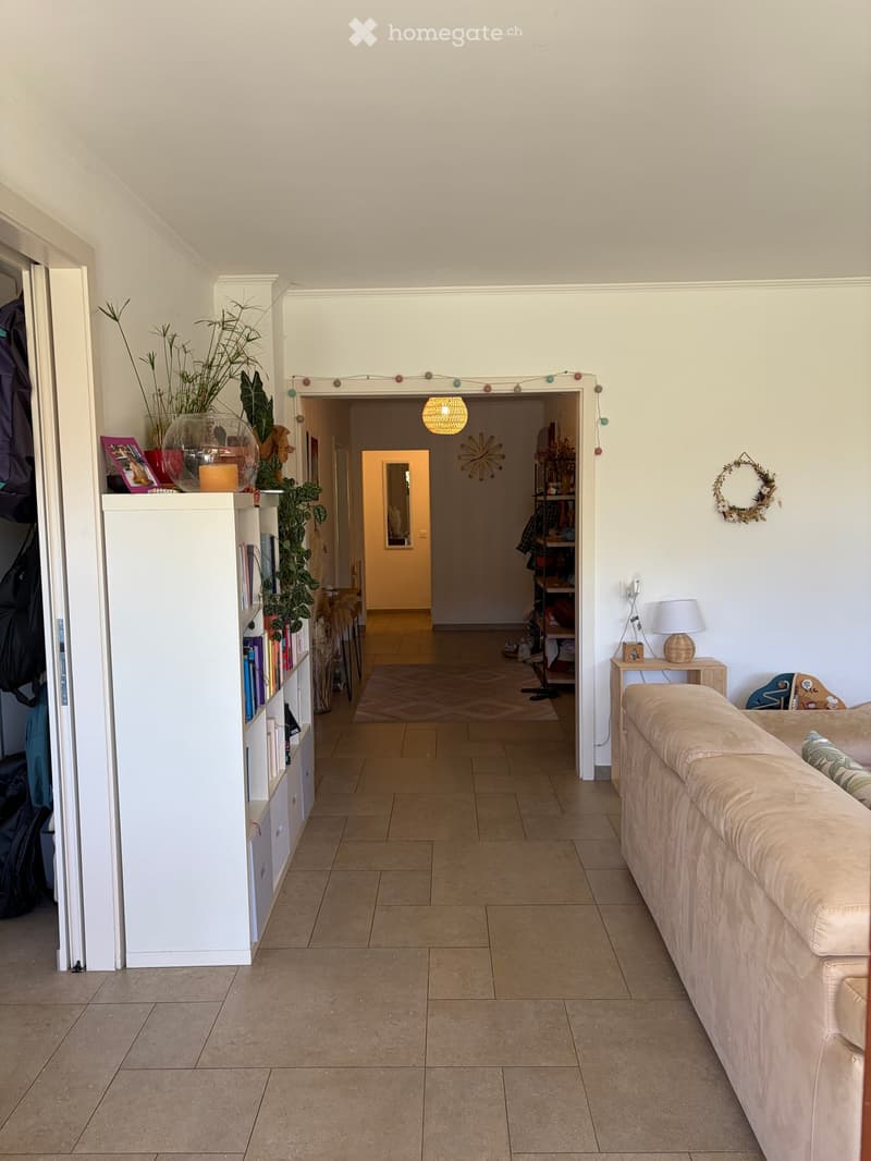A well-lit living room with a beige sofa, a small wooden table with a lamp, a bookshelf with books and plants, and a decorative wreath on the wall. The room has tiled flooring and leads to another room with a patterned rug.