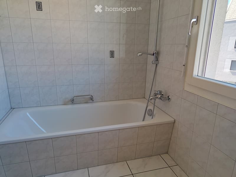 Modern bathroom with a bathtub, tiled walls, and a window. Features a handheld showerhead and a chrome faucet.