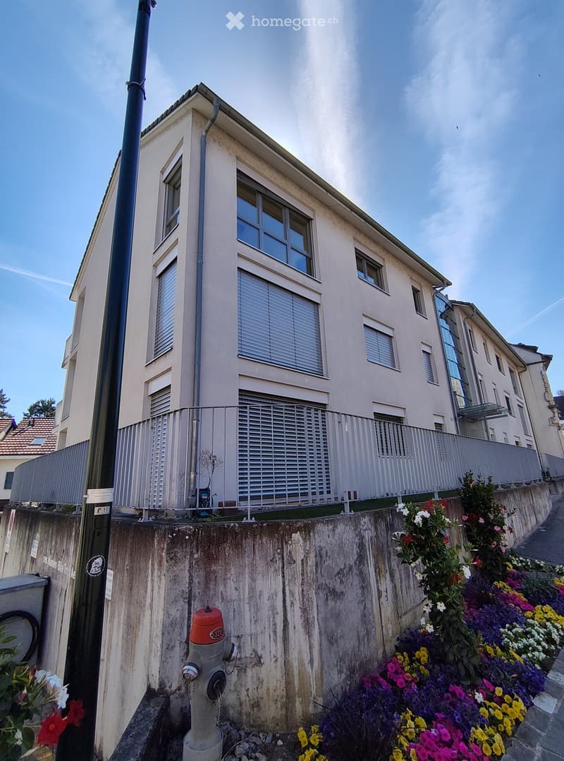 A two-story residential building with light-colored walls, multiple windows, and a metal fence along the top. The building is situated on a raised ground floor with a concrete retaining wall and colorful flower beds in front.
