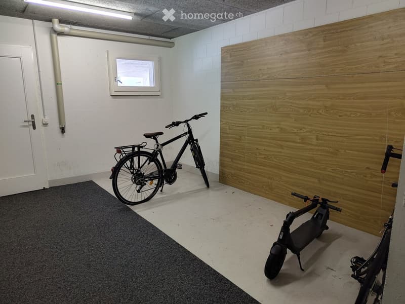 Empty room with white walls, a wooden panel accent wall, a small window, and a grey carpeted area. Contains a bicycle and an electric scooter.
