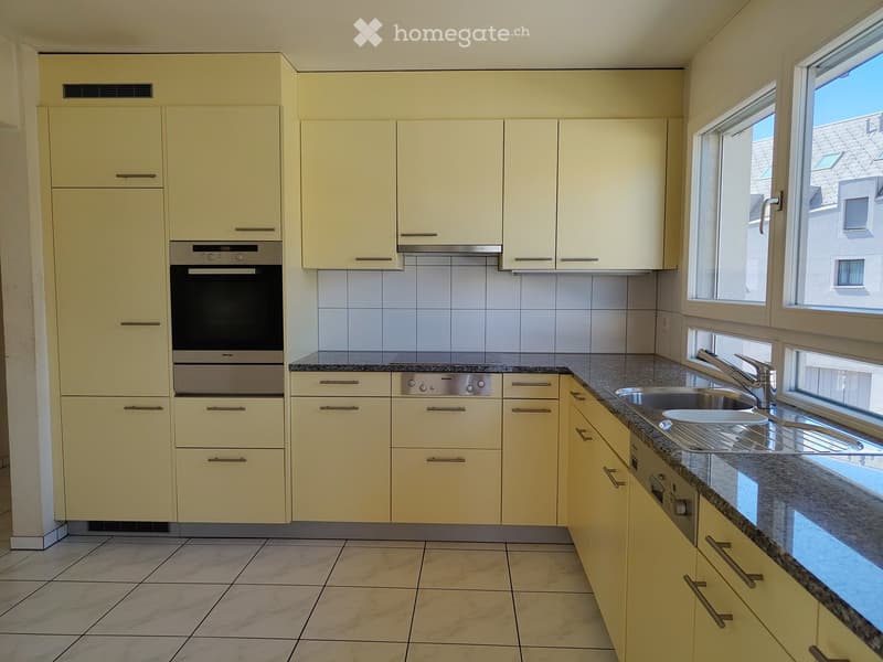 Modern kitchen with light-colored cabinets, granite countertops, and stainless steel appliances including an oven and dishwasher. The space features tiled flooring and large windows allowing natural light.