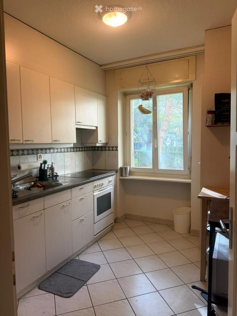 Bright kitchen with white cabinets, tiled floor, and a window overlooking a garden. Features include a sink, oven, countertop, and a small stool near the window.