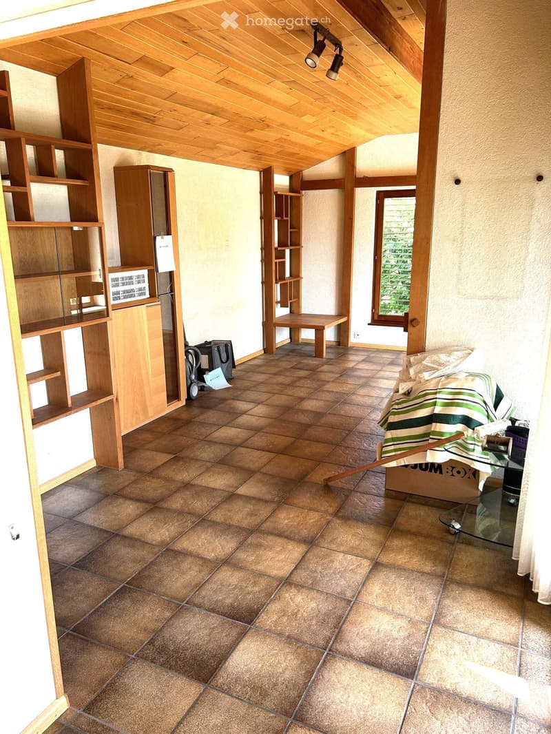 A furnished room with wooden shelves, a bench, a table, a chair, and a storage box. The room features tiled flooring and wooden beams on the ceiling.