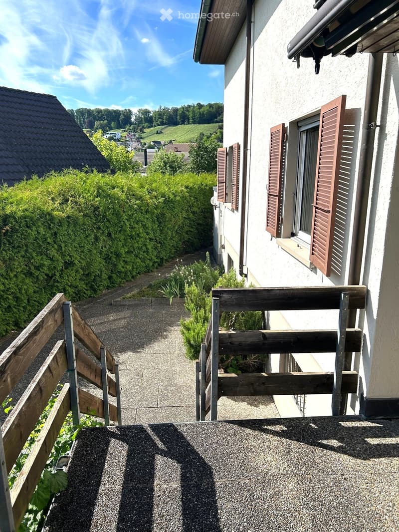 Balcony with wooden railing, gravel surface, and view of a house with shutters and surrounding greenery.