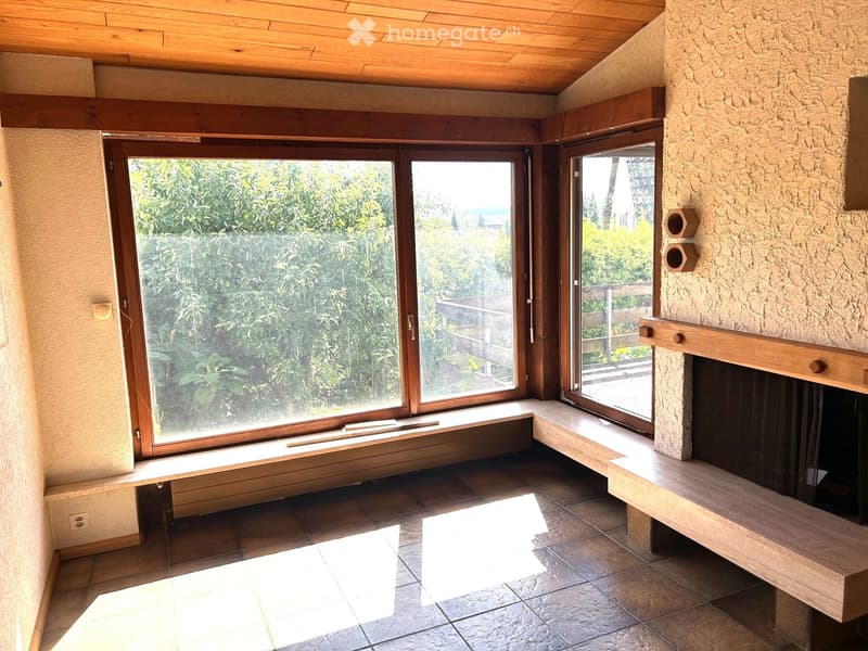 Living room with large windows offering a view of greenery, a stone fireplace, and wooden beams on the ceiling.