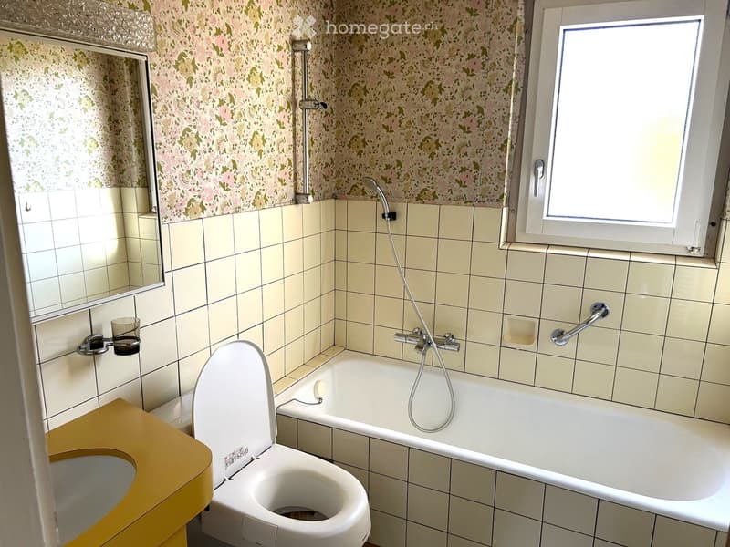 Bathroom with floral wallpaper, tiled walls, a toilet, a bathtub with a handheld shower, a mirror, and a small cup on the wall.