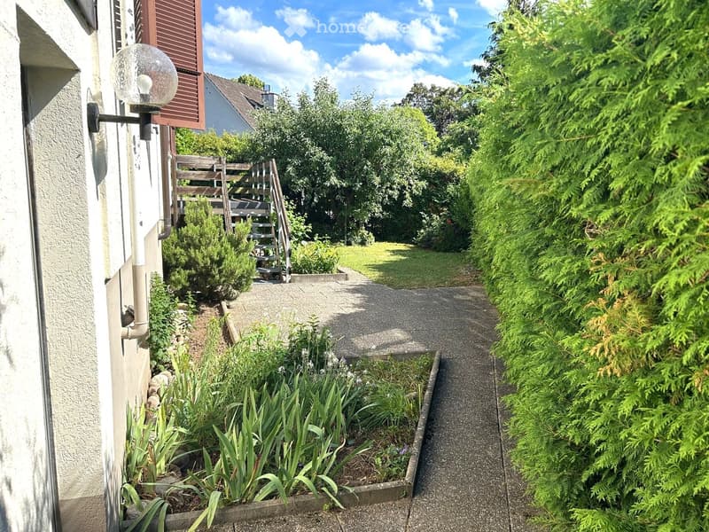 A narrow garden path lined with greenery and a wooden staircase leading to a raised area, with a wall-mounted light fixture on the left.